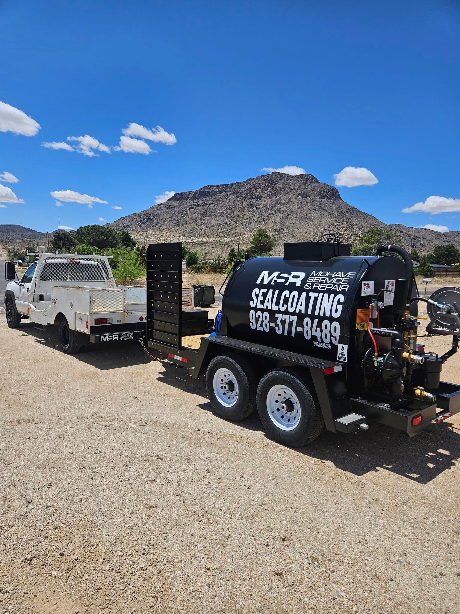 Black tow trailer with white lettering parked on gravel beside a pickup, desert mountain in the background.