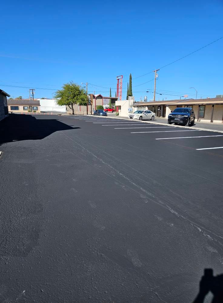 Empty parking lot with fresh black asphalt, a few cars, and storefronts under a clear blue sky