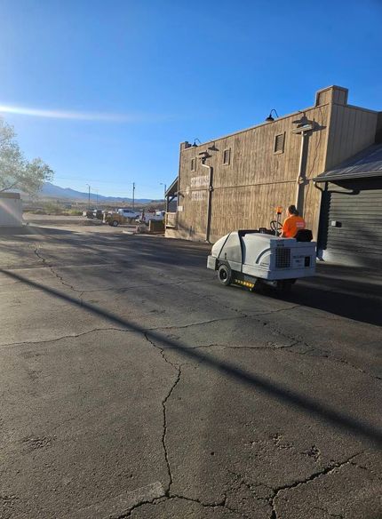 Worker in orange vest on a small street sweeper beside a stone building under a clear blue sky.