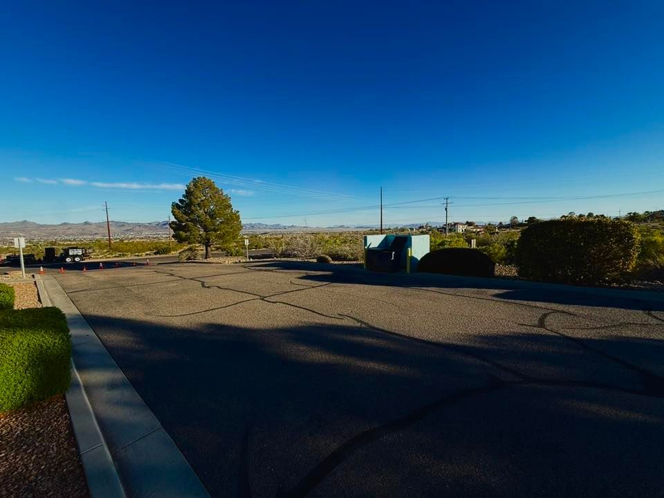 Wide empty parking lot under a clear blue sky with a tree, hedges, and distant mountains