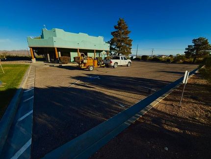 Wide rural roadside building with a green roof, parked vehicles, and a dirt lot under a clear blue sky