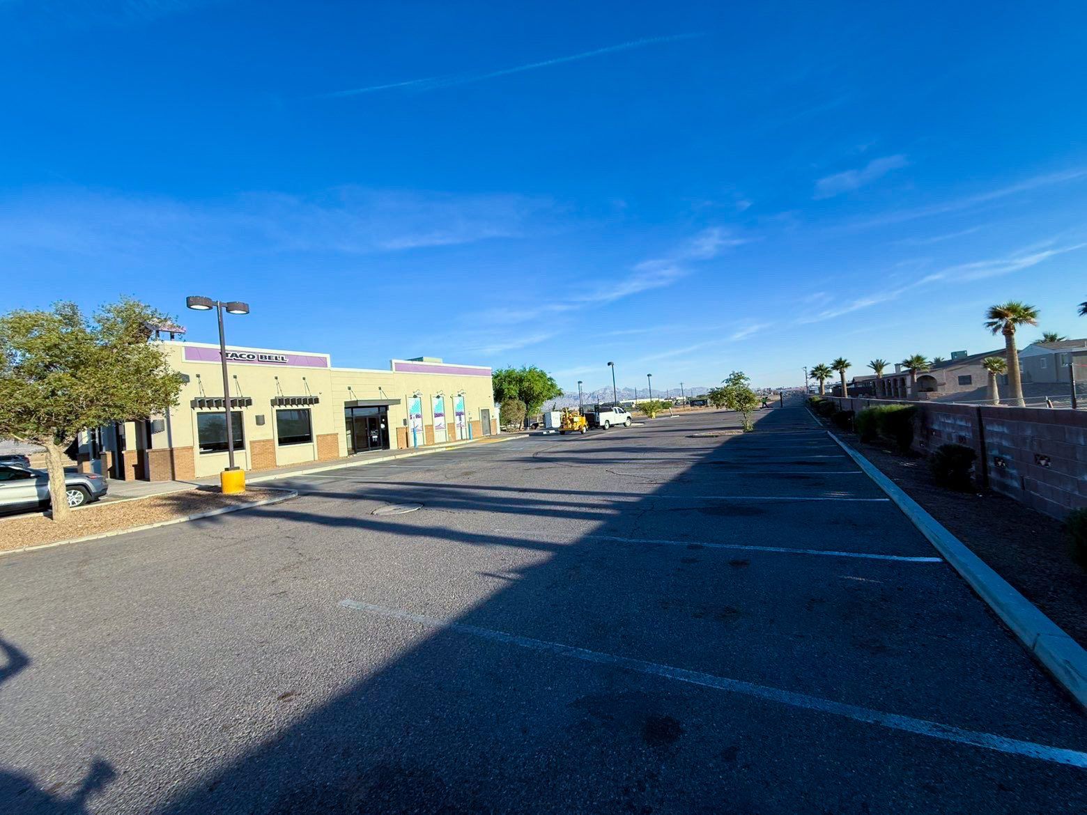Wide empty parking lot beside a beige building under a bright blue sky, with palm trees in the distance