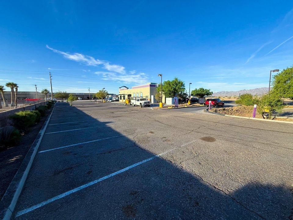 Empty gravel parking lot with blue sky, low buildings, and trees in the distance