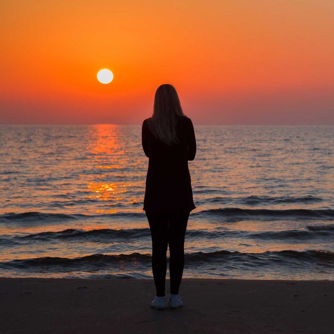 Woman standing on a beach, facing the setting sun over the ocean. Orange sky, dark silhouette.