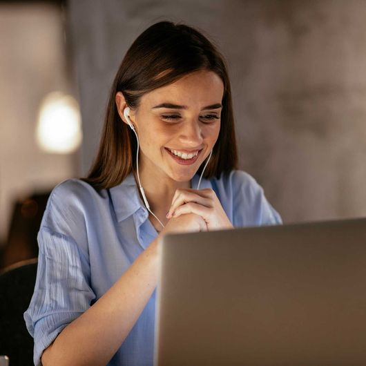 Woman smiling, wearing earbuds, looking at laptop screen in a dim room.
