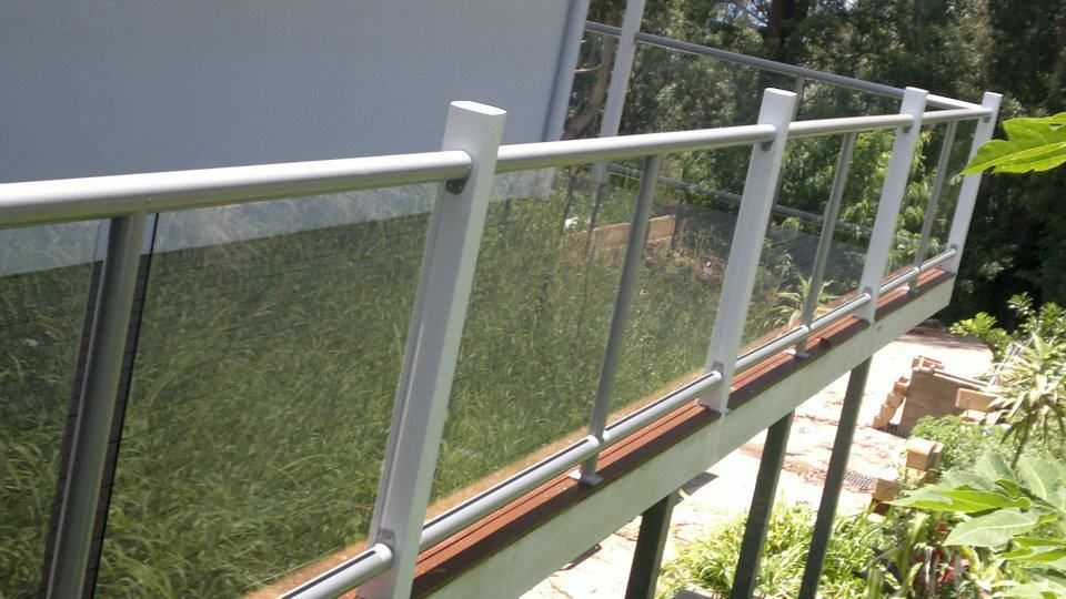 A Balcony With A Glass Railing Is Surrounded By Trees And Grass — Boambee Home Improvements in Coffs Harbour, NSW