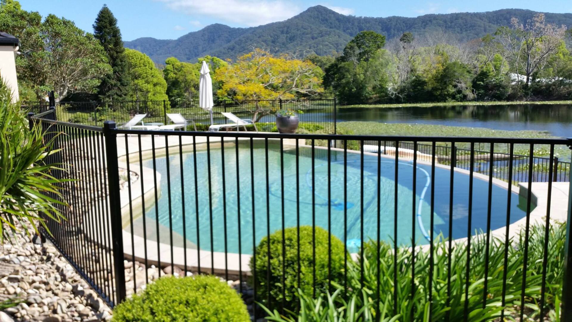 A Fence Surrounds A Large Swimming Pool With Mountains In The Background — Boambee Home Improvements in Coffs Harbour, NSW