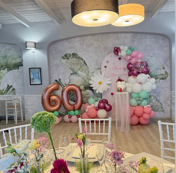 Allestimento per una festa di 60° compleanno con palloncini color oro rosa, composizioni floreali dai toni pastello e un arco di palloncini verdi e bianchi.