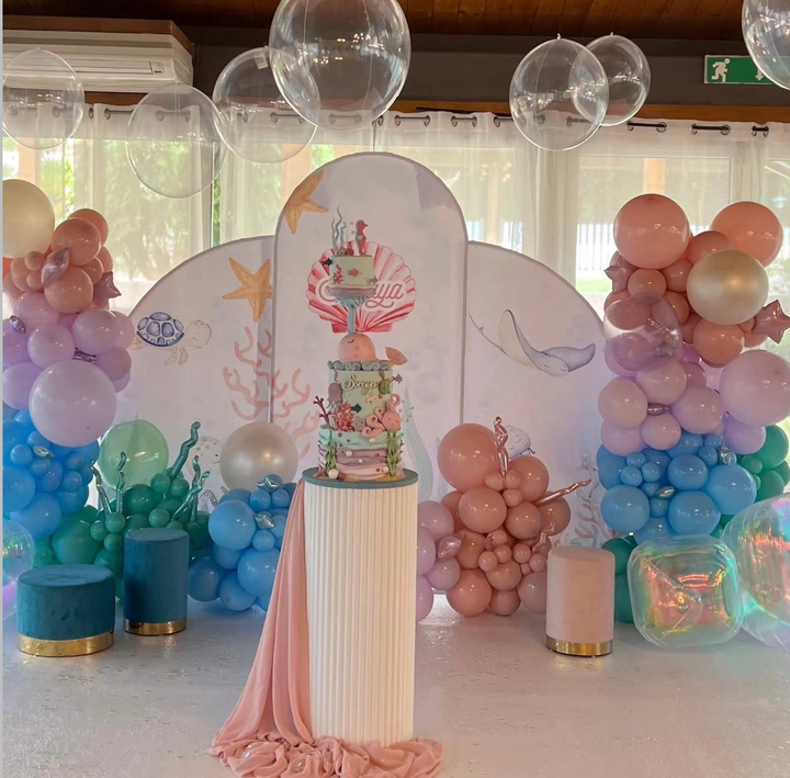 Sfondo per festa con palloncini dai colori pastello e torta floreale su un tavolo bianco con piedistallo.