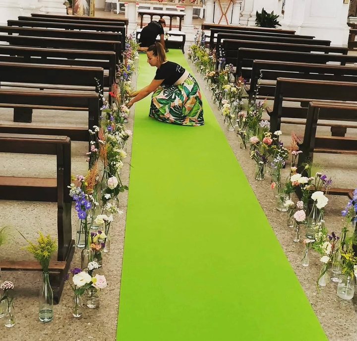Una donna inginocchiata su un corridoio verde brillante tra panchine ornate di fiori, sistema le decorazioni in una location addobbata.
