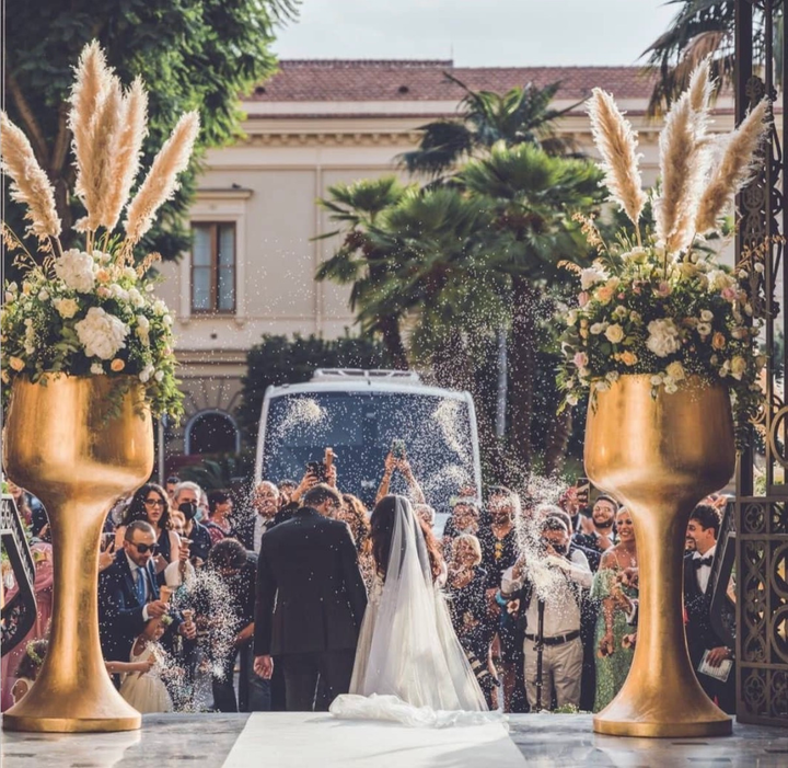 Gli sposi vengono rinfrescati con una spruzzata di champagne durante un matrimonio in giardino con gli invitati e vasi di fiori dorati.