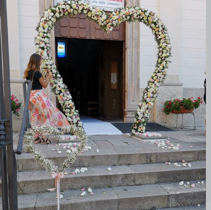 Un arco a forma di cuore ricoperto di fiori incornicia l'ingresso della chiesa, con una donna in piedi accanto e petali sui gradini.