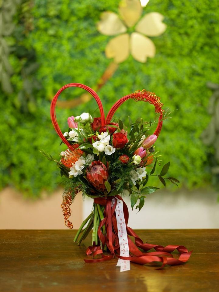 Composizione floreale a forma di cuore rosso con fiori rossi, bianchi e rosa su un tavolo, su uno sfondo floreale verde.