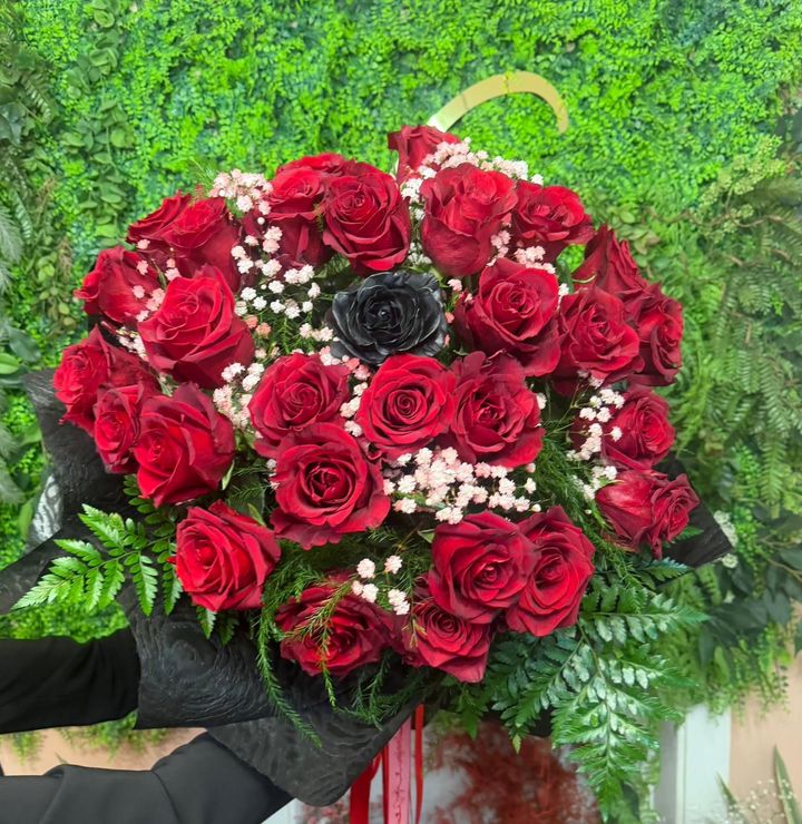 Un bouquet composto da numerose rose rosse e una singola rosa nera, impreziosito da piccoli fiori bianchi e verde, su uno sfondo verde.