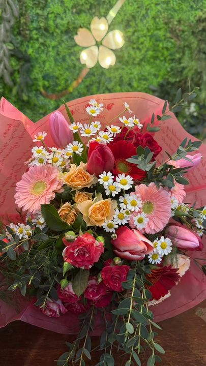 Bouquet colorato di fiori misti in carta rosa con fogliame verde, disposto su una superficie di legno.