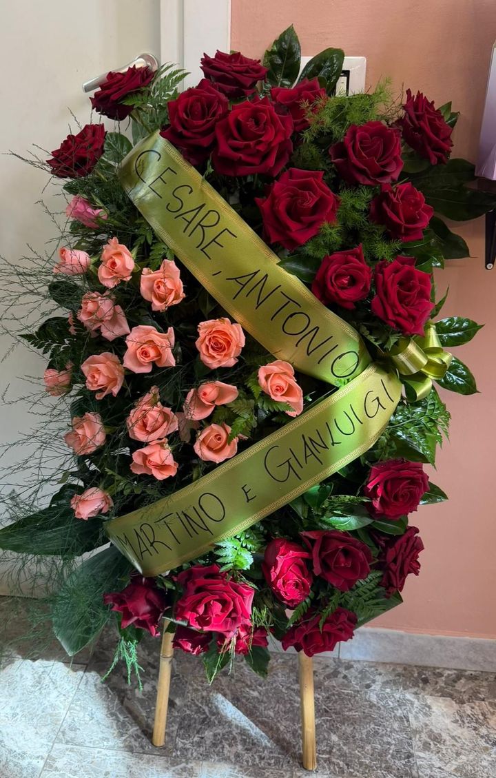 Una composizione floreale funebre alta, con rose rosso scuro e rosa tenue, e un nastro su cui sono incisi i nomi in italiano.