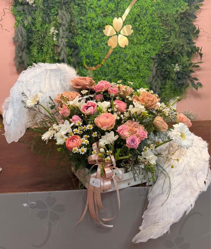 Composizione floreale rosa e color pesca con drappeggio di tessuto bianco su un tavolo, contro una parete verde di piante.