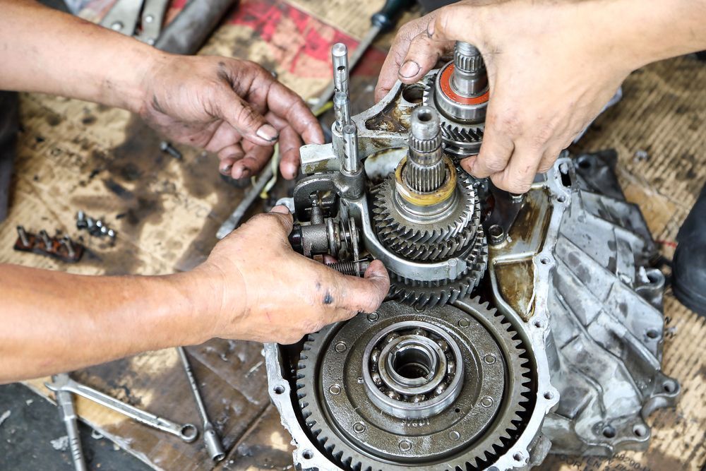 Hands repairing a disassembled gear transmission on a dirty workbench with tools and oil-stained parts
