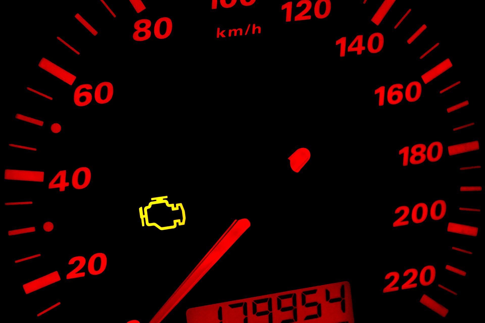 Car dashboard speedometer at 0 km/h with red-lit dial and engine warning light on