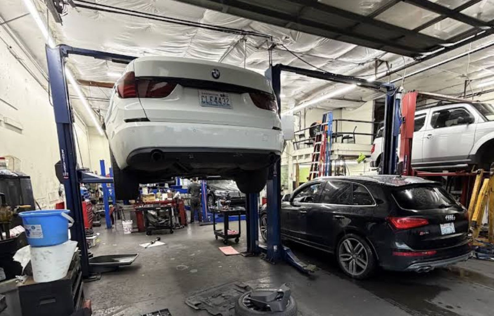 Auto repair shop with a white sedan lifted on a hoist and a black sedan parked below, tools and equipment around.