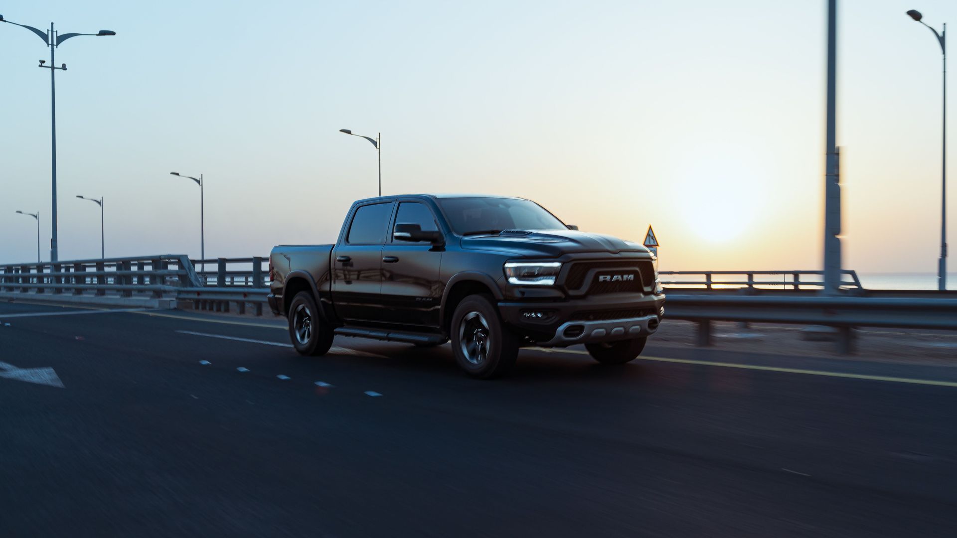 Black pickup truck driving on a bridge at dusk with streetlights and calm water in the background