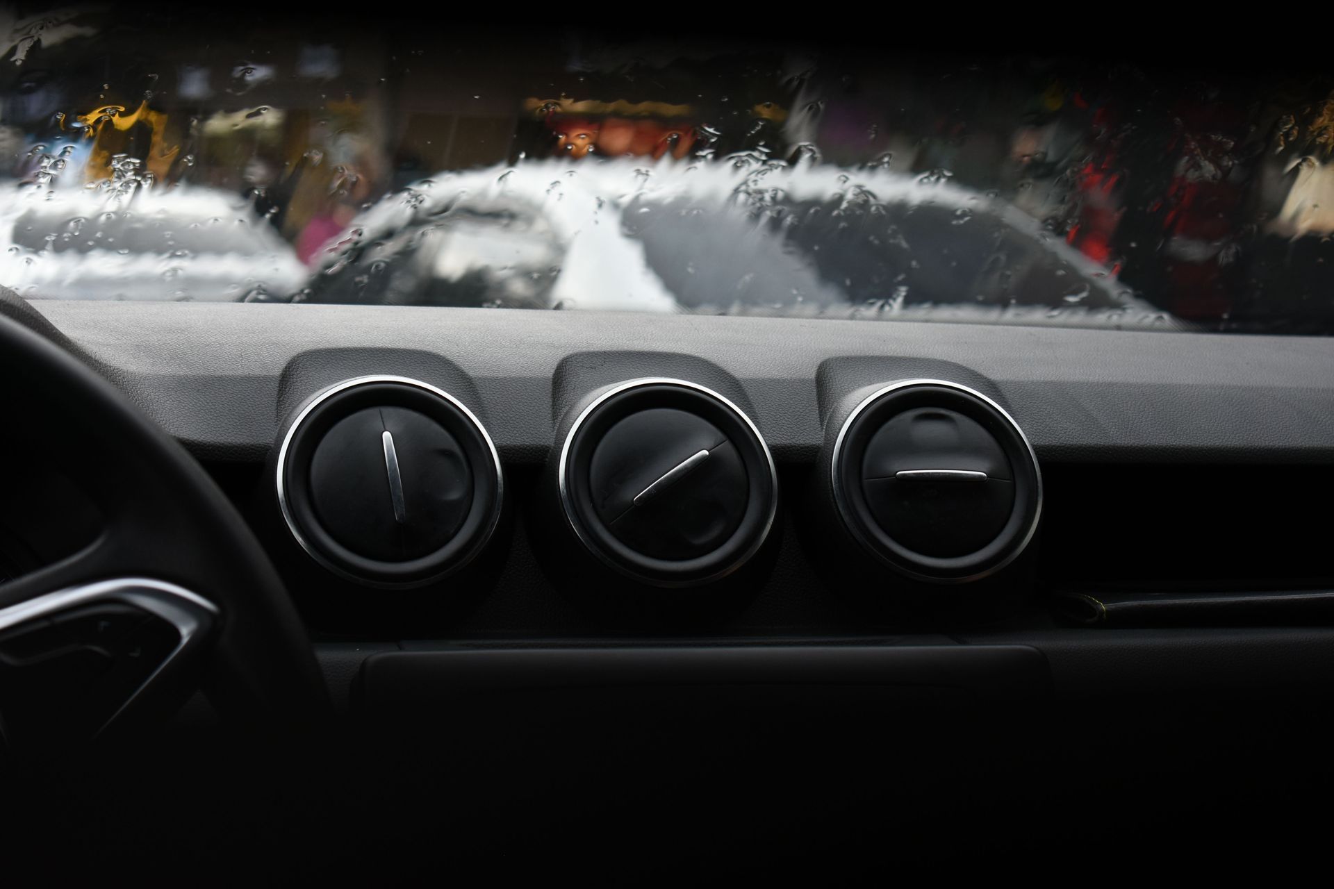 Car dashboard with three circular air vents, looking through a rain-covered windshield at a white car ahead
