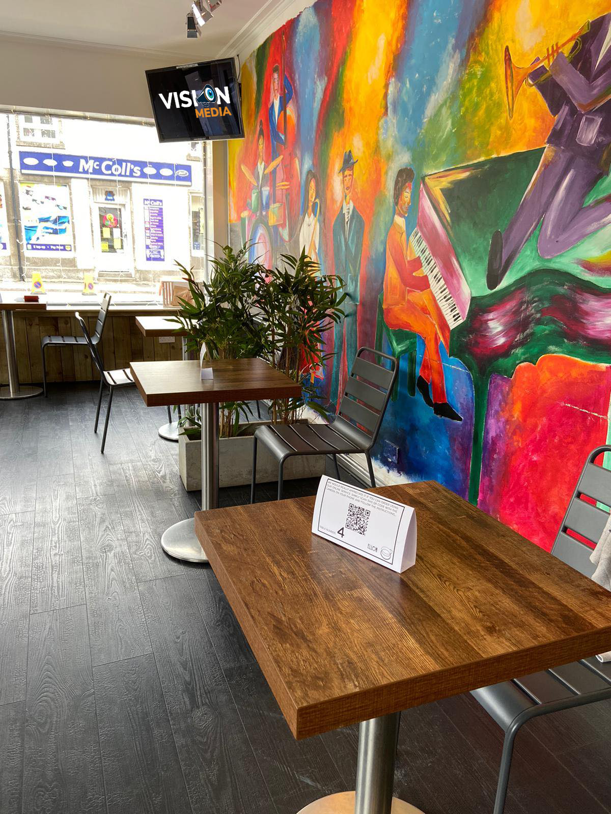 Café seating area with wooden tables, chairs, potted plants, and a bright colorful mural on the wall