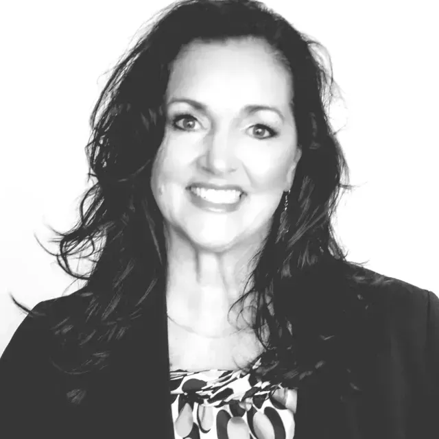 Woman smiling, wearing a black blazer and patterned blouse, in a professional setting.