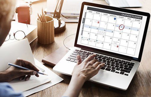 Person using a laptop to view a calendar planner, writing on a notepad at a desk.