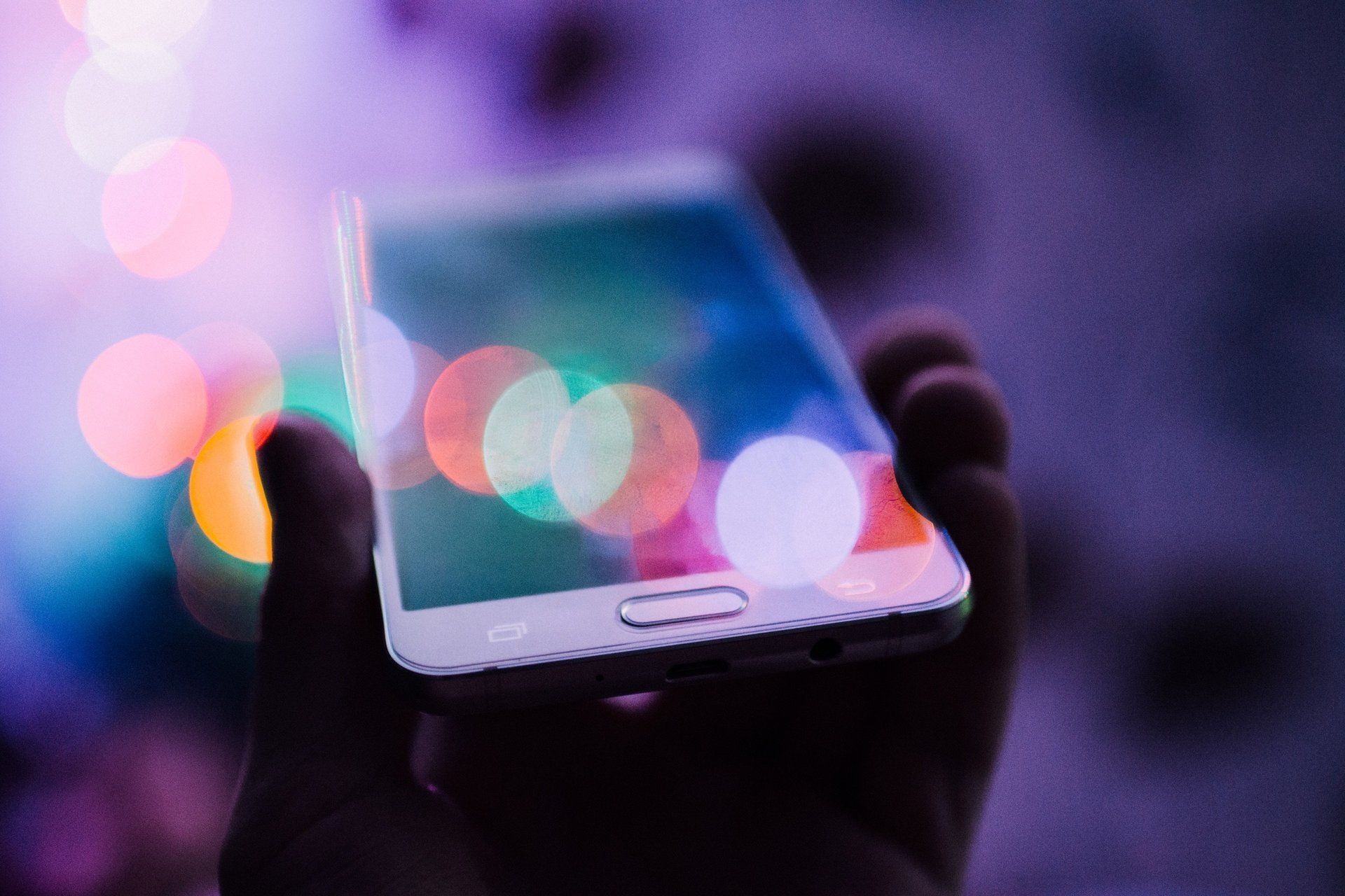 Hand holding a smartphone reflecting colorful bokeh lights in a dim environment.