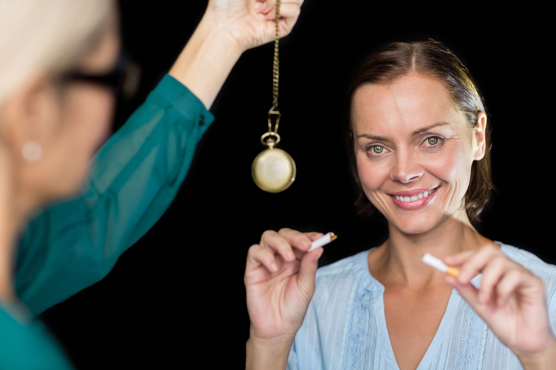 Woman breaking a cigarette, smiling, with hypnotist and swinging pocket watch in background.
