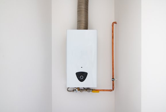 White wall-mounted water heater with copper pipe and exhaust duct, in the corner of a room.