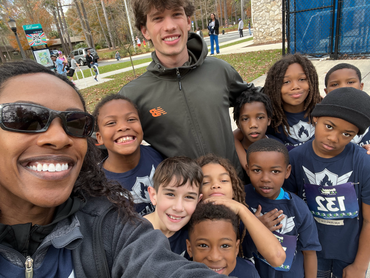 Group selfie with two adults and a diverse group of smiling children outdoors. They are in front of a park.