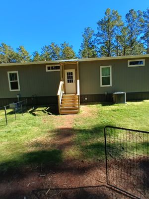 Single-story green house with a front porch and steps, set on a grassy yard with trees behind.