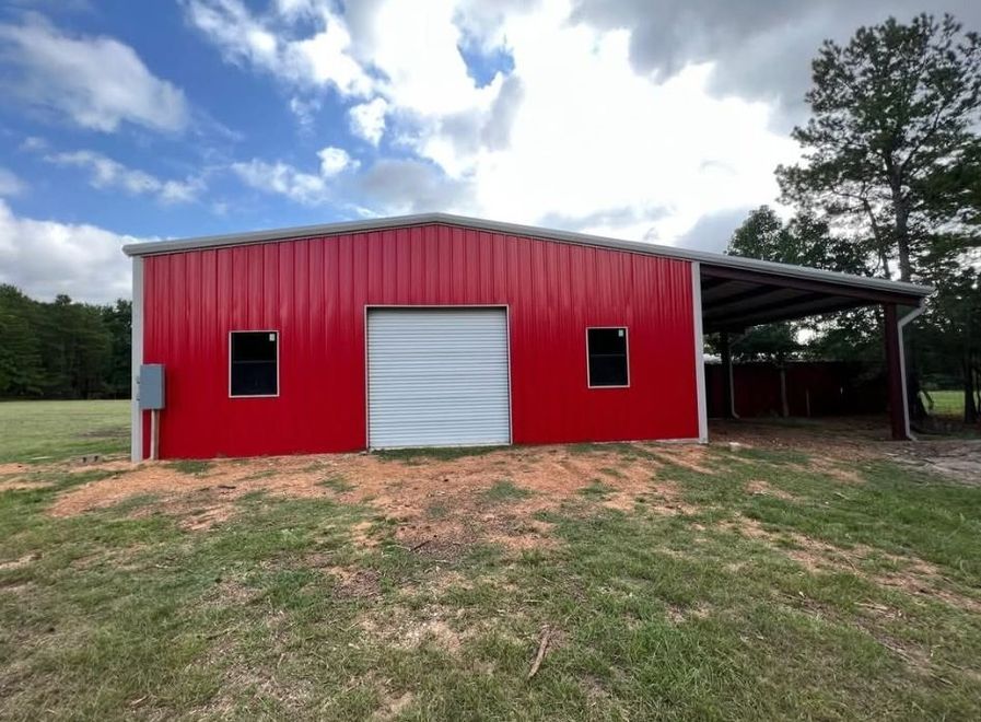 Red metal barn with white roll-up door and black windows in a grassy field under cloudy sky