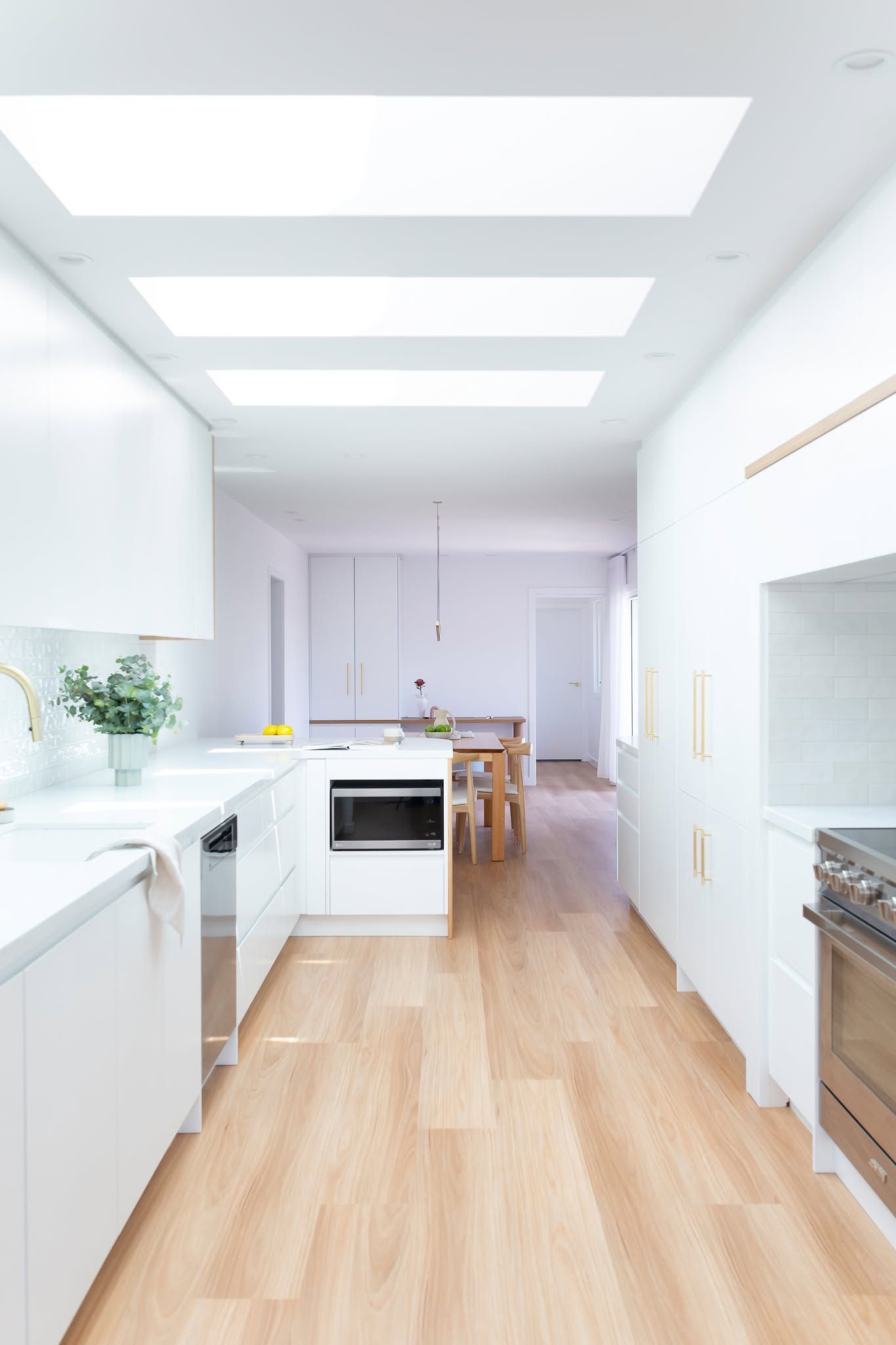 Bright modern kitchen with white cabinets, wood floors, skylights, and a dining area in the distance — Complete Property Projects In Phillip, ACT