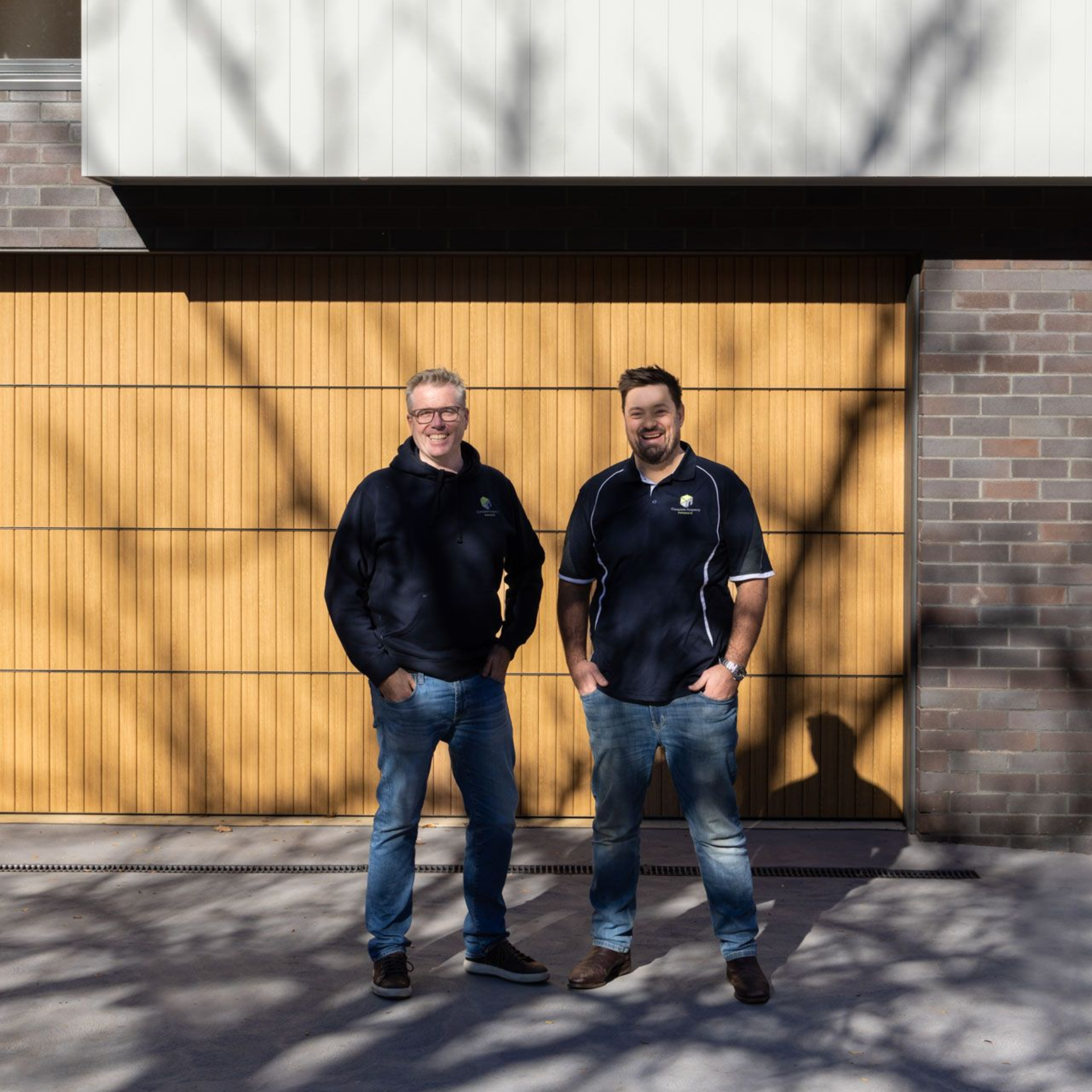 Two men standing in front of a large yellow garage door and brick building, casting long shadows. — Complete Property Projects In Phillip, ACT