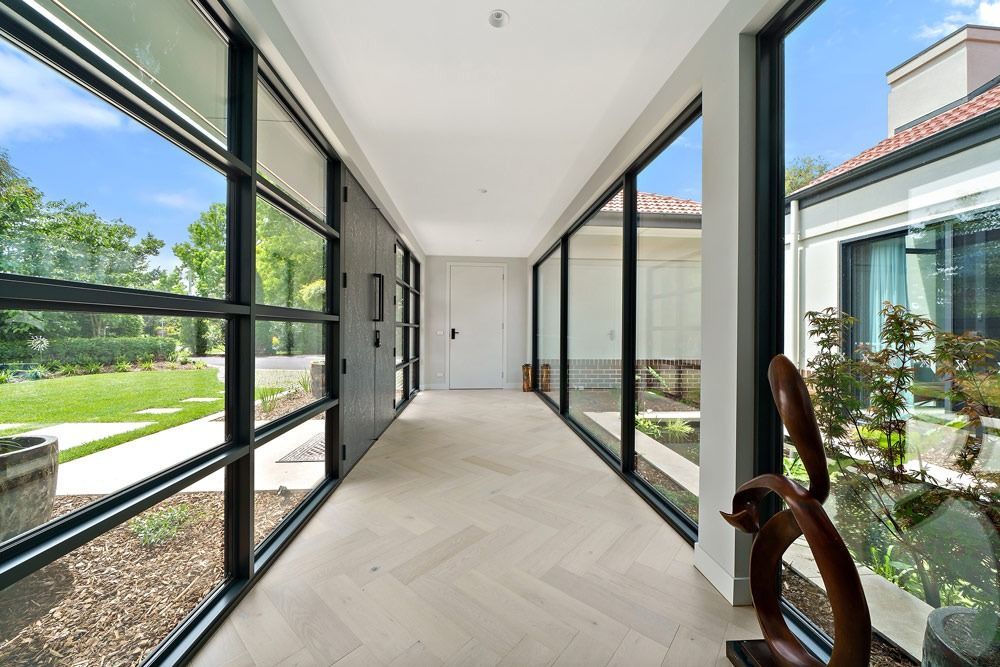 Bright modern hallway with large glass windows, white walls, and herringbone flooring, opening to greenery outside