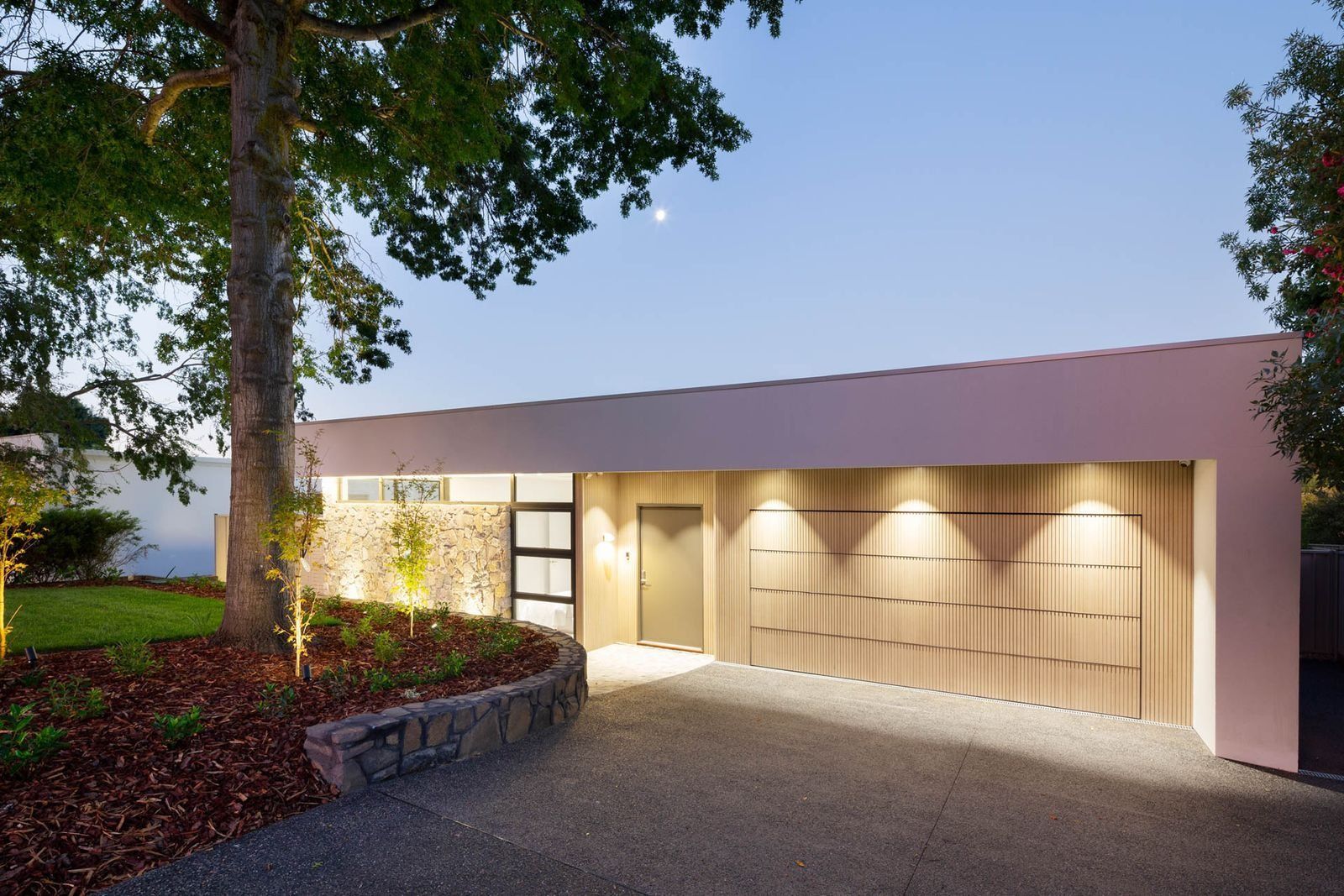 Modern House Exterior with A Lit Garage, Stone Wall, and Trees at Dusk — Complete Property Projects In Phillip, ACT