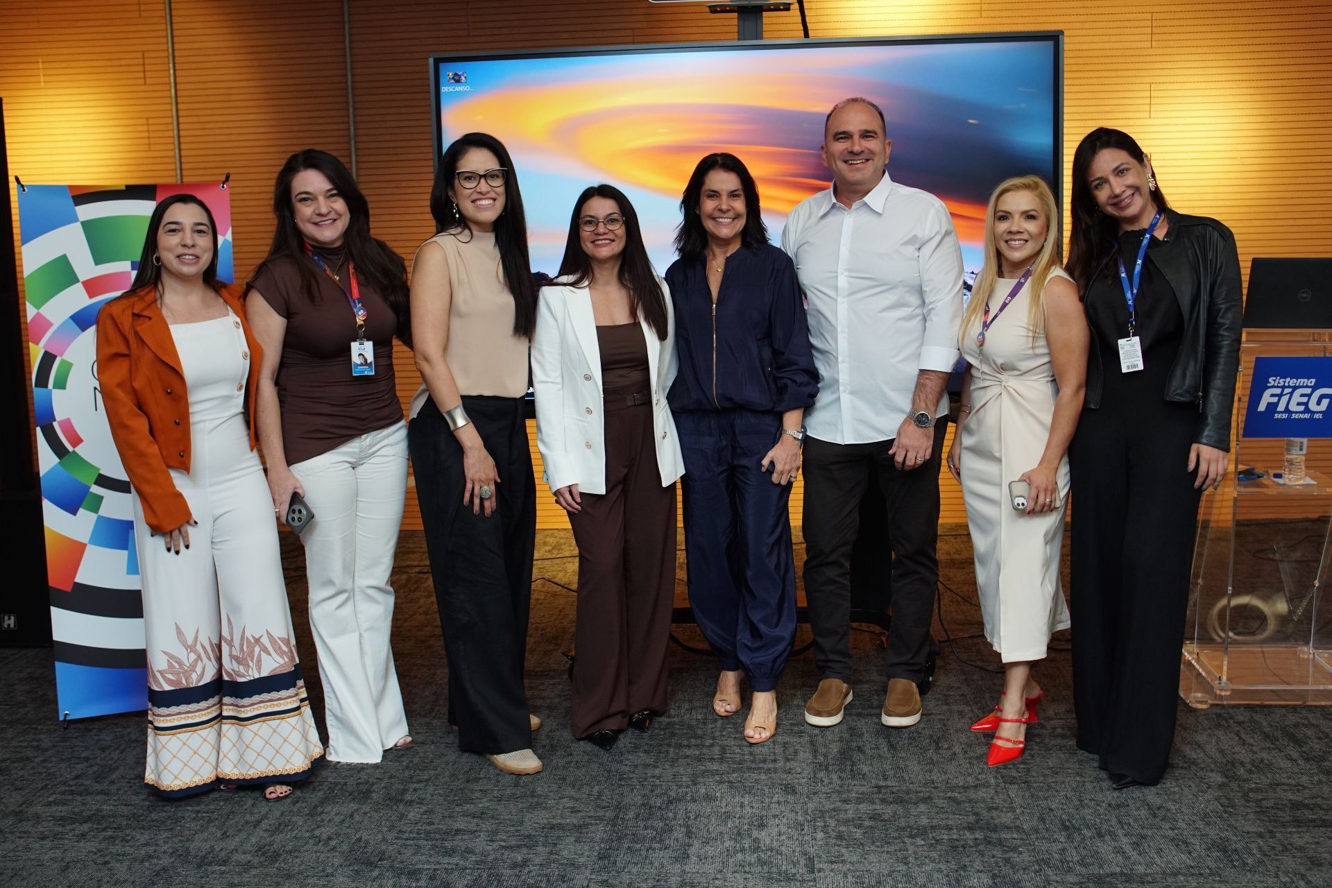 Sete pessoas posando em um estande de conferência com um colorido pôr do sol ao fundo.