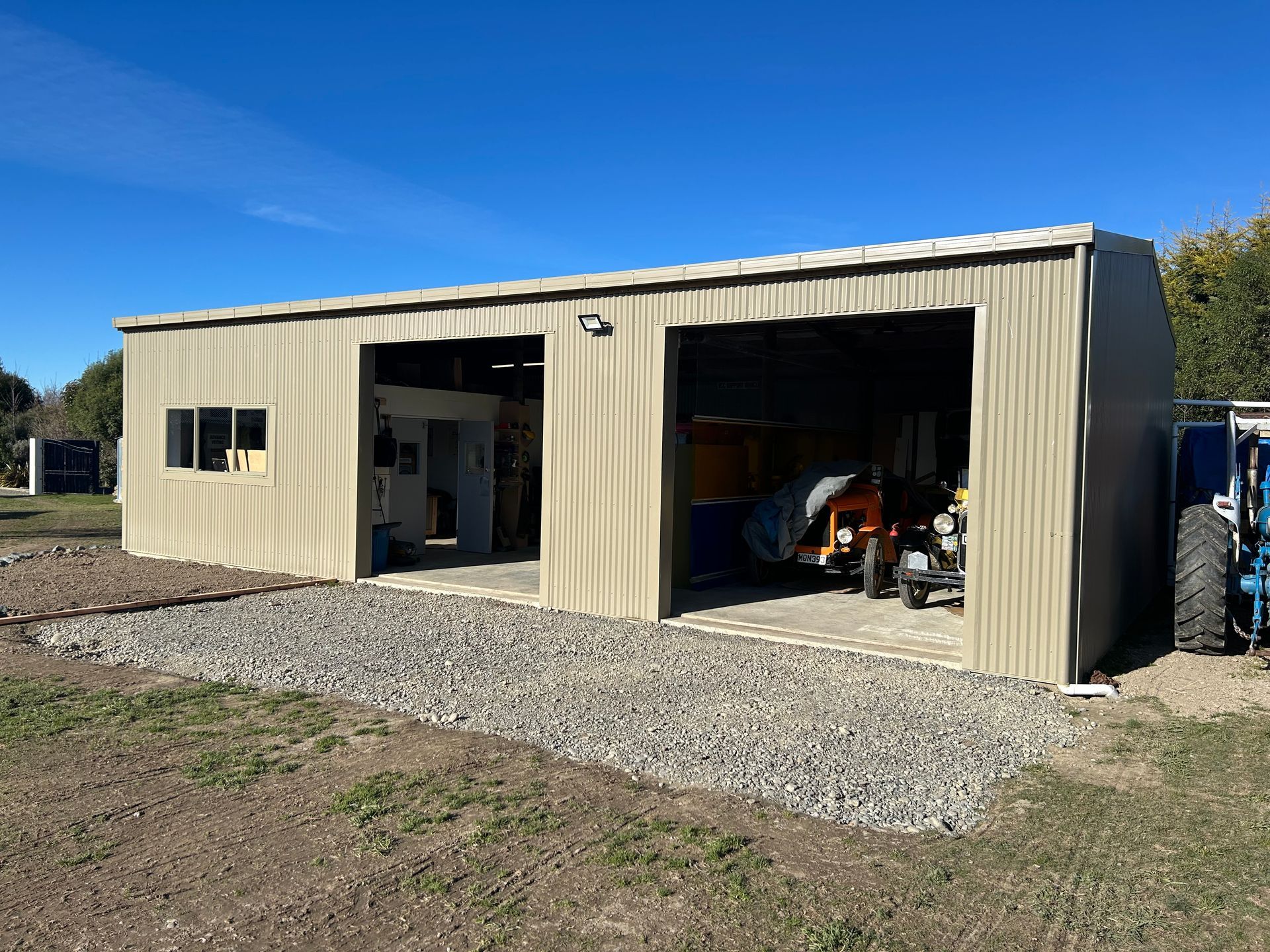 Tan metal garage with two open bays, gravel base, blue sky.