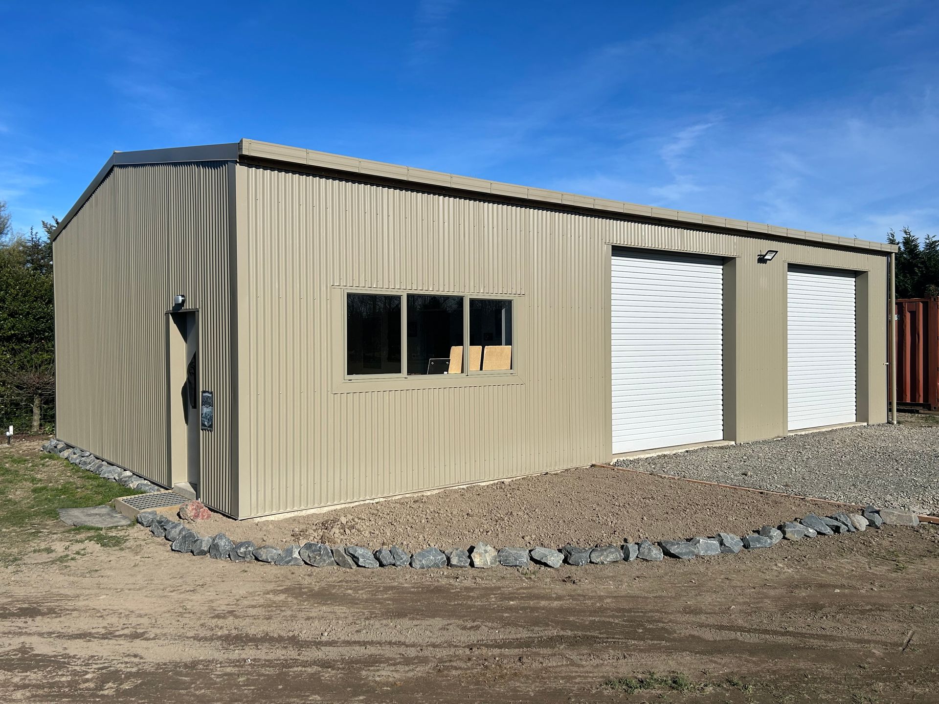 Tan metal shed with two garage doors, window, and rock border against blue sky.