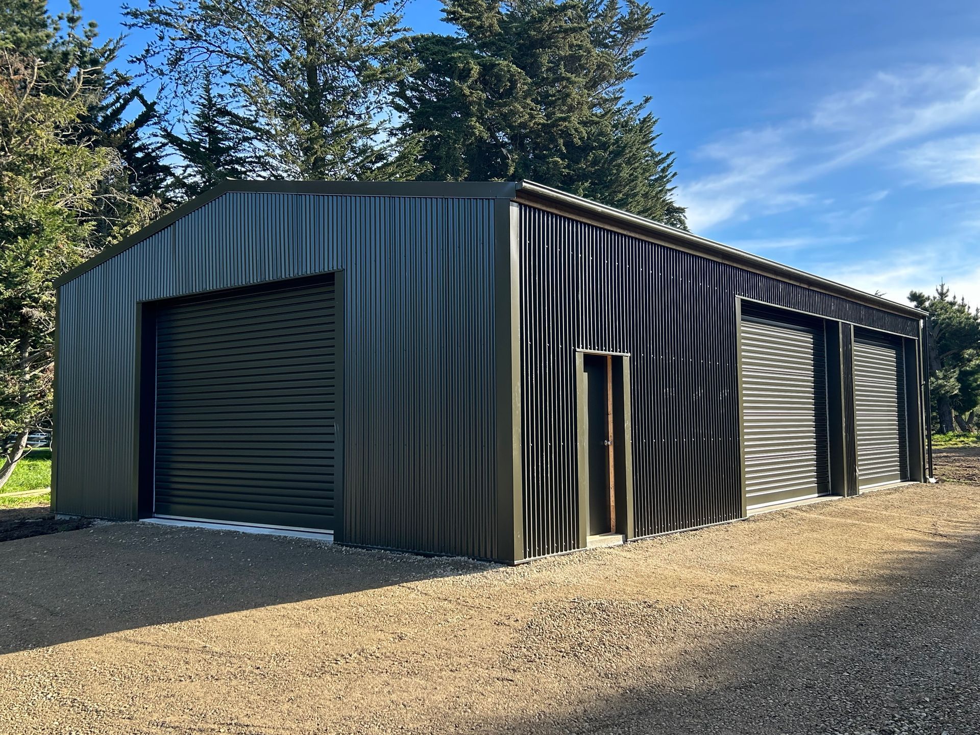 Dark green corrugated steel building with three garage doors on a gravel lot under a blue sky.