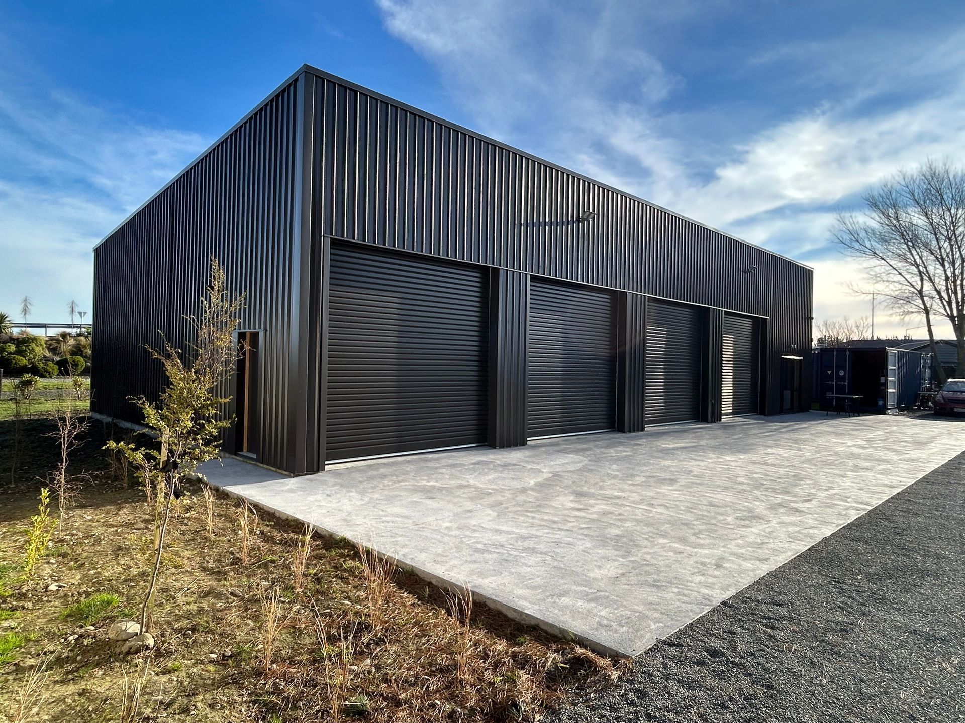 Black building with three garage doors on concrete pad, against a blue sky.