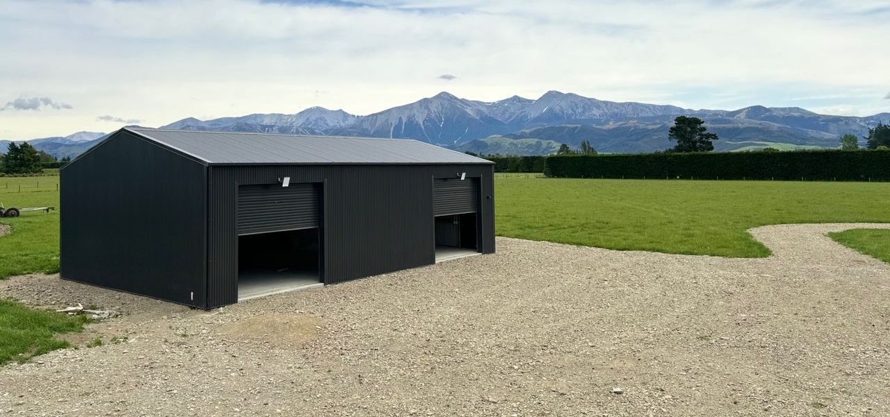 A black shed is sitting in the middle of a grassy field with mountains in the background.