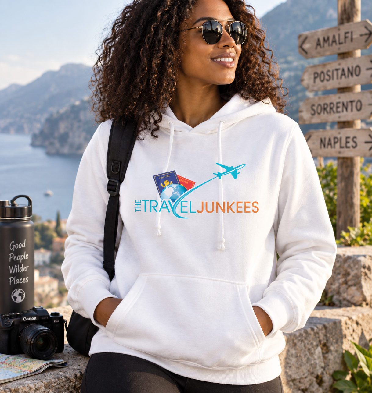Woman in black hoodie at a scenic coastal overlook, with mountains, sea, and a travel sign in the background