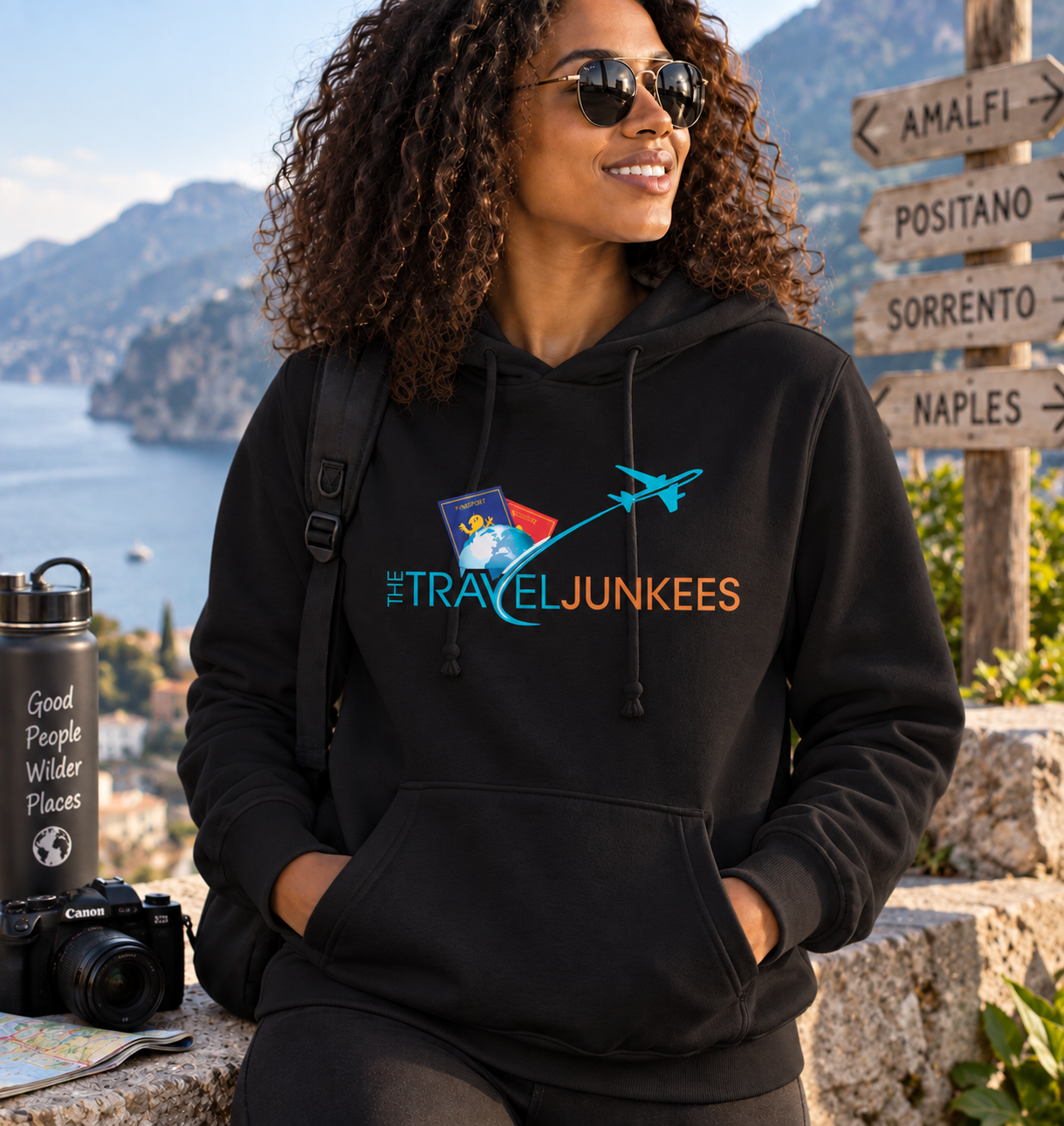 Woman in black hoodie at a scenic coastal overlook, with mountains, sea, and a travel sign in the background