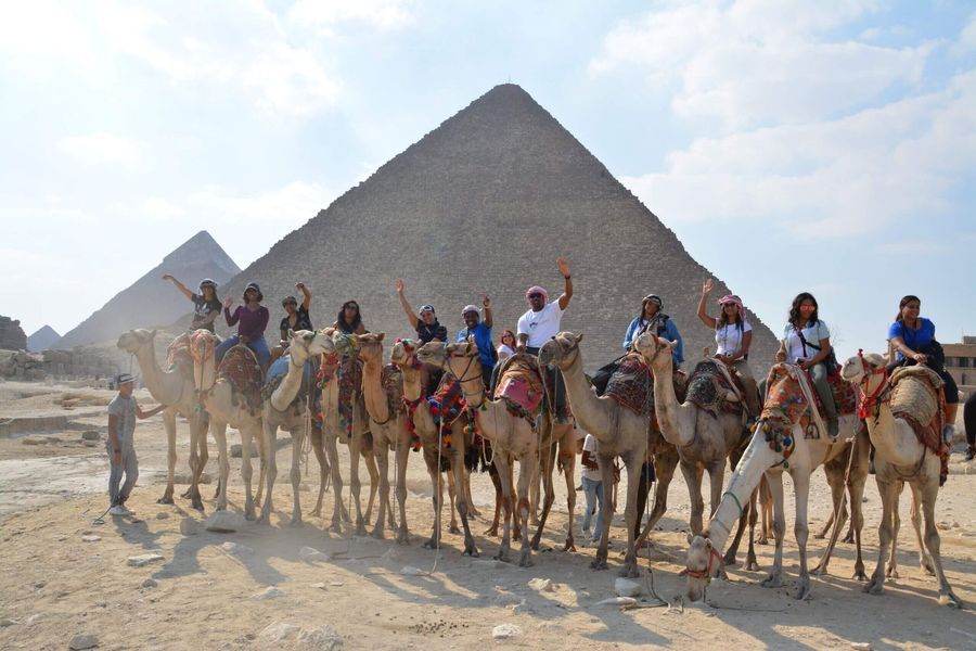Tourists riding camels in front of the pyramids in the desert under a bright sky