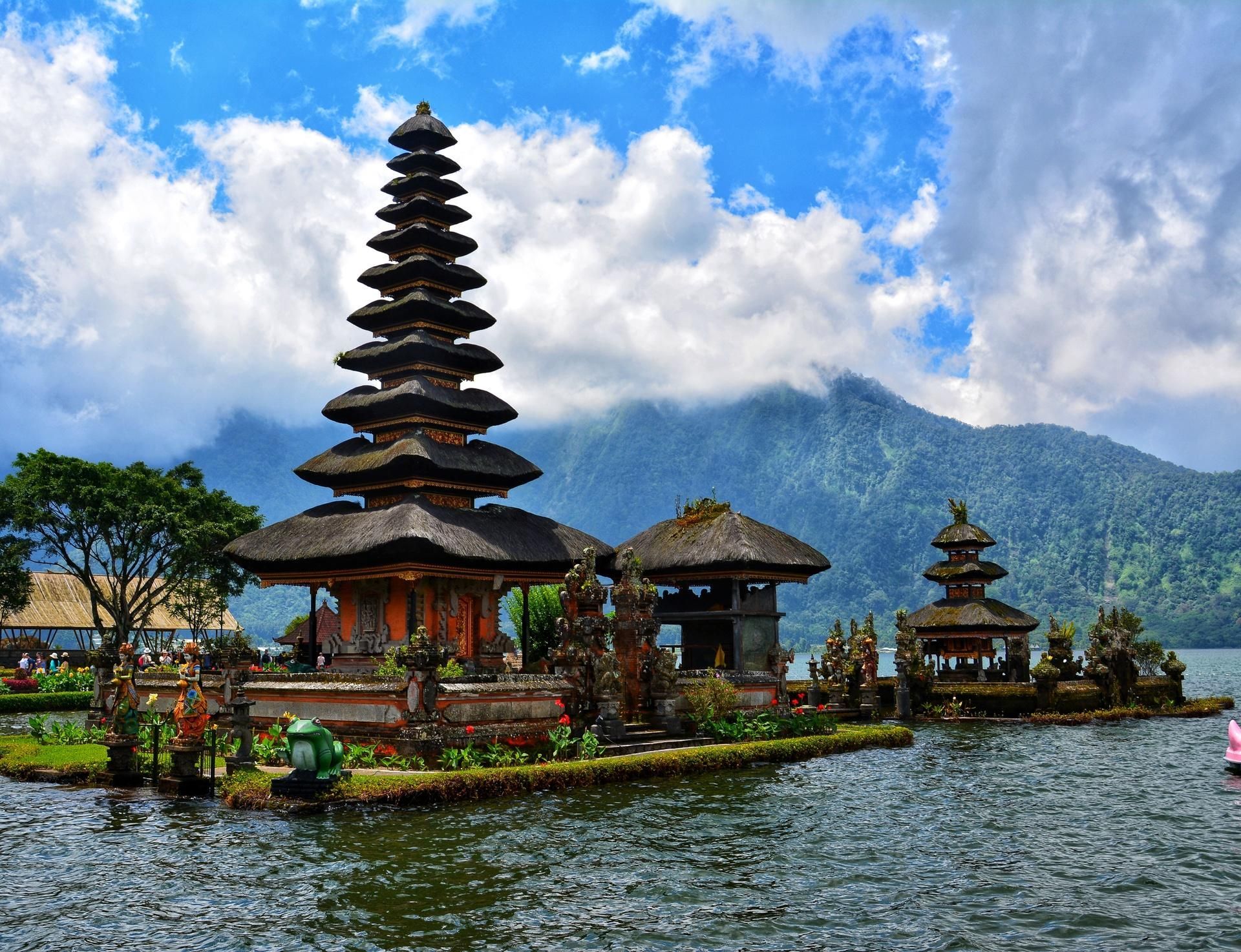 Balinese temple pavilion and tall tiered shrine on a lake, with mountains and clouds in the background