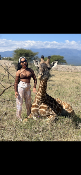 Woman stands beside a giraffe sculpture in a grassy field with mountains in the background.